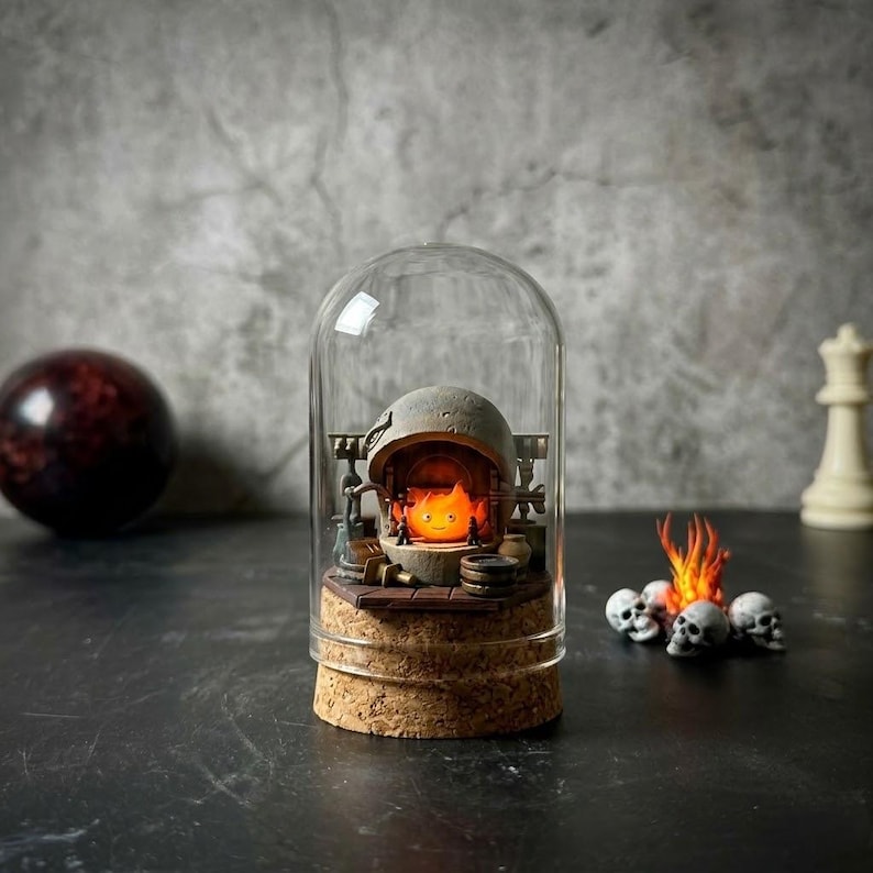 Pu&ograve; includere: Una cupola di vetro con base in sughero contenente un focolare in pietra grigia in miniatura con un personaggio a forma di fiamma arancione brillante all'interno. Accanto alla cupola si trova un piccolo mucchio decorativo di teschi bianchi.