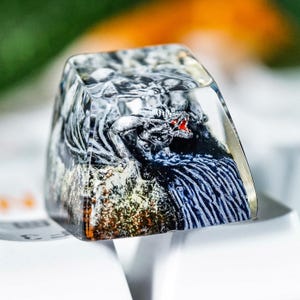 Puede incluir: Una tecla personalizada con una escultura de dragón dentro de resina transparente. El dragón es gris con detalles rojos, sobre un fondo rocoso texturizado. La tecla está diseñada para encajar en un teclado mecánico.