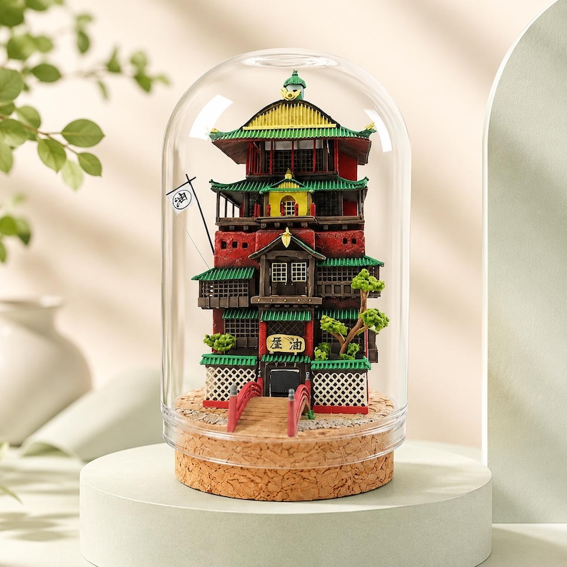 K&ouml;nnte beinhalten: Ein Miniatur-Geb&auml;udemodell mit mehreren Stockwerken unter einer Glaskuppel auf einem Korksockel. Das Bauwerk hat rote W&auml;nde, gr&uuml;ne Ziegeld&auml;cher, eine kleine rote Br&uuml;cke, eine wei&szlig;e Flagge mit dem schwarzen Kanji f&uuml;r &Ouml;l und kleine gr&uuml;ne B&auml;ume. &Uuml;ber dem Eingang befindet sich ein Schild mit dem Kanji f&uuml;r &Ouml;l.