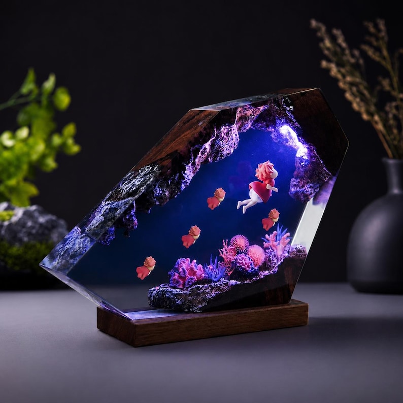 May include: A decorative lamp featuring a dark wood frame encasing a clear resin block. Inside, a blue-lit underwater scene displays a small figure in a red dress swimming among pink coral and several small orange fish figures on a wooden base.