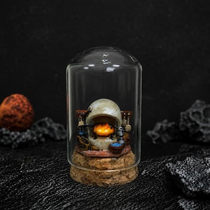 Pu&ograve; includere: Un diorama in miniatura di un focolare all'interno di una campana di vetro con base in sughero. La scena presenta un piccolo personaggio a forma di fiamma arancione in un camino color pietra, circondato da utensili, una pentola blu e un supporto.
