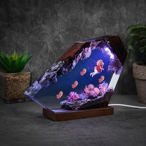 May include: A decorative lamp featuring a dark wood frame enclosing a clear resin block. Inside the resin, a miniature scene depicts a figure in a red dress swimming among small orange creatures, purple coral, and pink sea anemones with blue light.