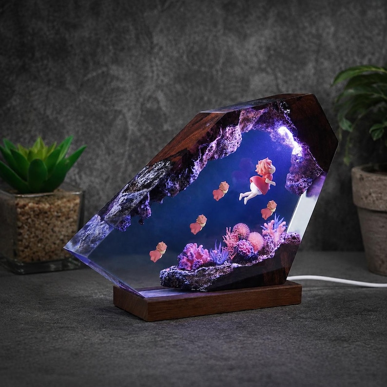 May include: A decorative lamp featuring a dark wood frame enclosing a clear resin block. Inside the resin, a miniature scene depicts a figure in a red dress swimming among small orange creatures, purple coral, and pink sea anemones with blue light.