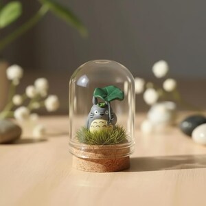 May include: A small gray creature with a cream-colored belly and whiskers stands on a patch of green grass inside a clear glass dome with a cork base. The creature holds a large green leaf over its head like an umbrella, featuring dark eyes.