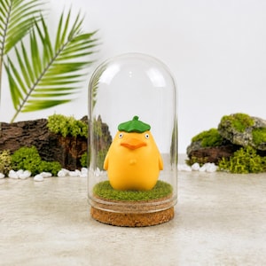 May include: A yellow figurine of a duck-like character wearing a green hat, displayed inside a glass cloche. The figurine sits on a bed of green moss atop a cork base. Decorative elements include moss-covered rocks and greenery.