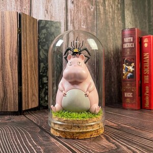 Puede incluir: Una figura de hipop&oacute;tamo rosa con vientre gris y una ara&ntilde;a negra encima, encerrada en una c&uacute;pula de cristal. La figura se asienta sobre una base de corcho con c&eacute;sped artificial. Art&iacute;culo decorativo.