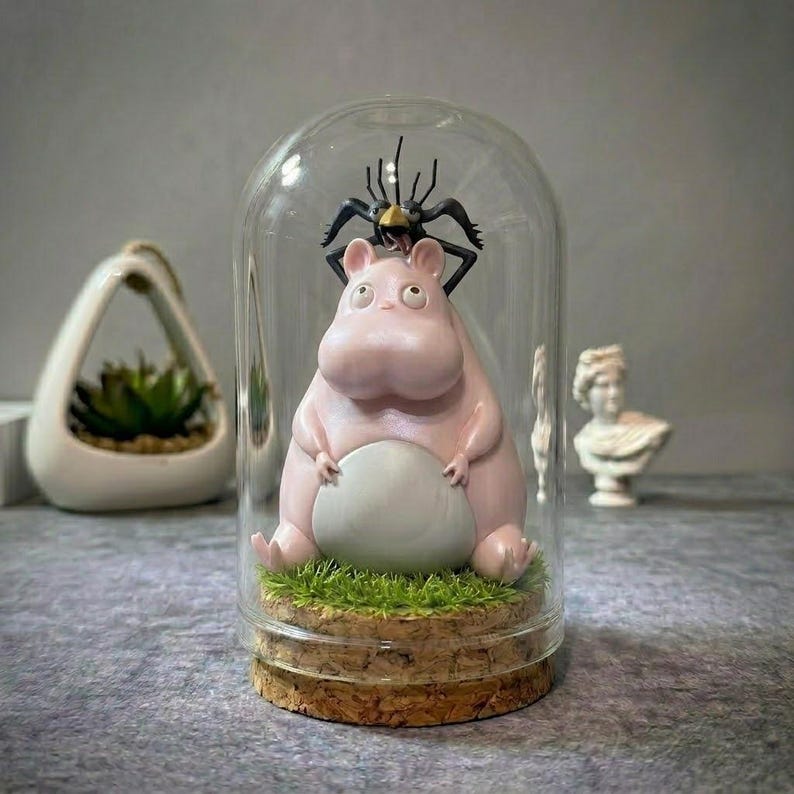 Puede incluir: Una campana de cristal decorativa caprichosa con una figura de hipop&oacute;tamo rosa con vientre gris y una figura de ara&ntilde;a negra en la parte superior. El hipop&oacute;tamo est&aacute; sentado sobre un lecho de c&eacute;sped artificial verde sobre una base de corcho. Un peque&ntilde;o macetero blanco y un busto est&aacute;n en el fondo.