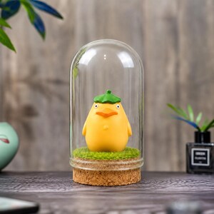 May include: A yellow figurine of a bird-like character wearing a green hat, encased in a glass dome. The figurine sits on a bed of green faux grass, with a cork base. The background is a blurred wooden surface.