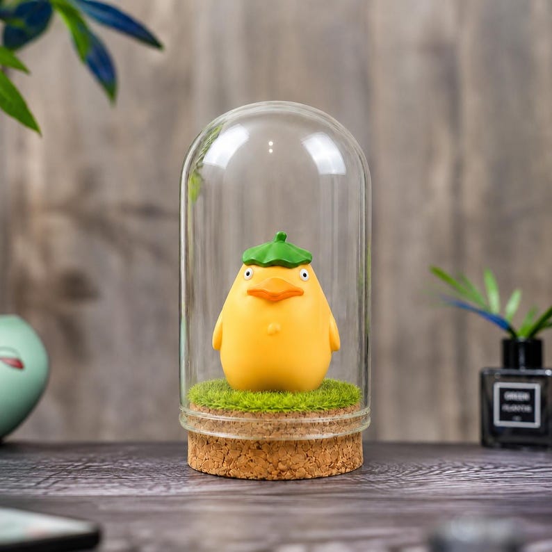 May include: A yellow figurine of a bird-like character wearing a green hat, encased in a glass dome. The figurine sits on a bed of green faux grass, with a cork base. The background is a blurred wooden surface.