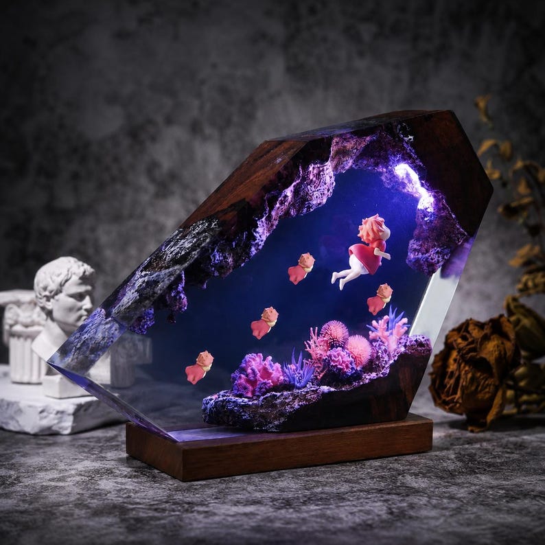 May include: A decorative lamp featuring a dark wood frame enclosing a clear resin block with an underwater scene. Inside, a small figure in a red dress swims among pink and purple coral formations, illuminated by a bright blue internal light.