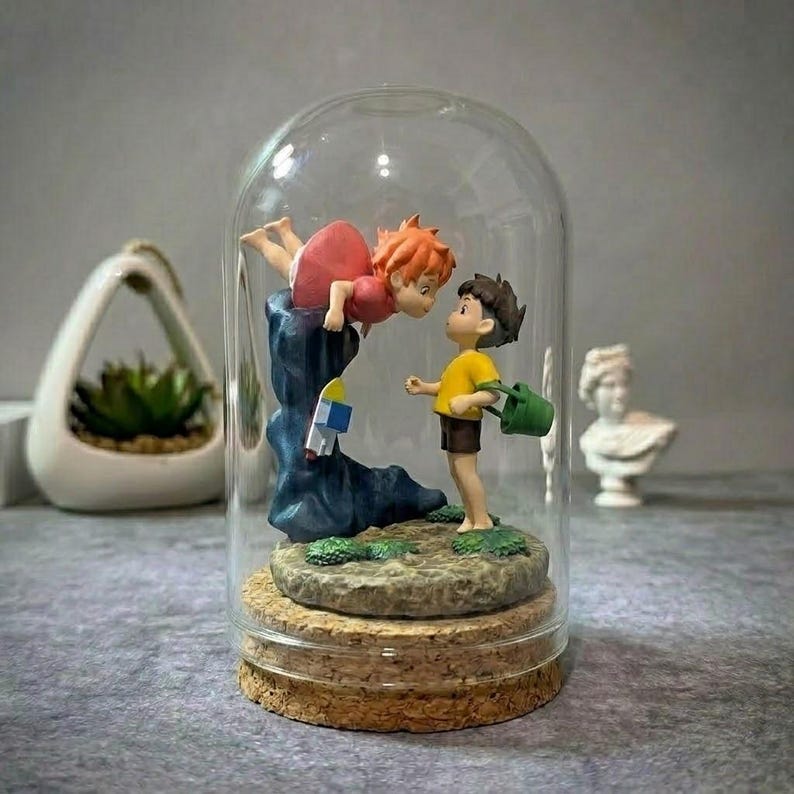 May include: A glass dome display featuring two small figurines on a cork base. One figure has orange hair and a red dress, positioned above a dark blue wave. The other figure wears a yellow shirt and brown shorts, holding a small green bucket.