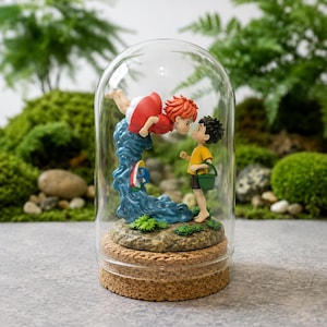 May include: A glass dome display featuring a figurine of a red-haired character in a red dress leaping from a blue wave toward a dark-haired character in a yellow shirt holding a green bucket. The figures stand on a textured base atop a cork stand.