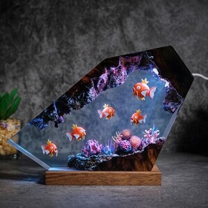 May include: A decorative resin lamp featuring orange fish swimming in a simulated underwater scene with coral and rock formations. The lamp has a wooden base and a geometric shape, with a light shining from within.