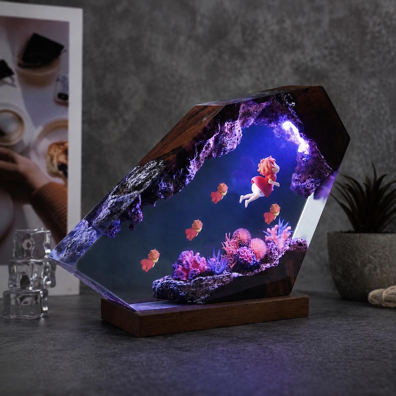 May include: A decorative lamp featuring a dark wood base and a resin block containing a miniature underwater scene. Inside the clear resin, a small figure with orange hair and a red dress swims among several tiny orange fish and colourful coral.