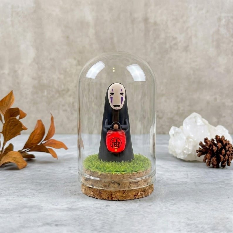 Op de afbeelding: Een figuurtje van een in het zwart gekleed personage met een wit masker dat een kleine rode lantaarn vasthoudt met het zwarte kanji-teken voor olie erop gedrukt. Het figuurtje staat op een stukje kunstgras in een glazen stolp met een kurken voet.