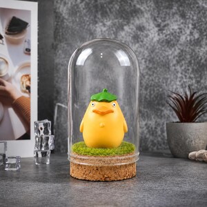 May include: A yellow figurine of a duck-like creature with a green hat, displayed under a glass dome. The figurine sits on a bed of green faux grass, atop a cork base. The background is a neutral gray.