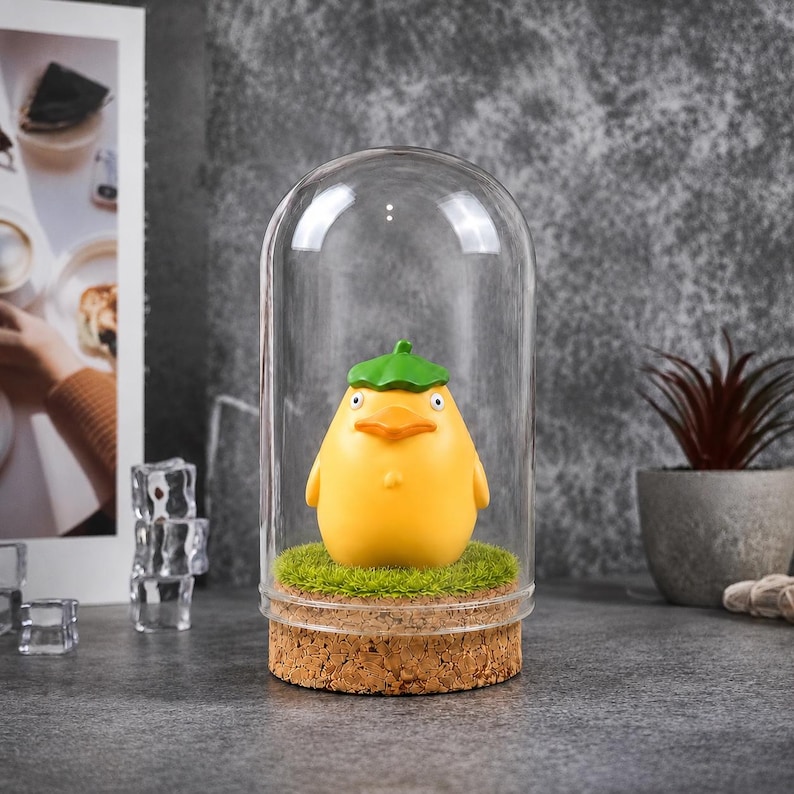 May include: A yellow figurine of a duck-like creature with a green hat, displayed under a glass dome. The figurine sits on a bed of green faux grass, atop a cork base. The background is a neutral gray.