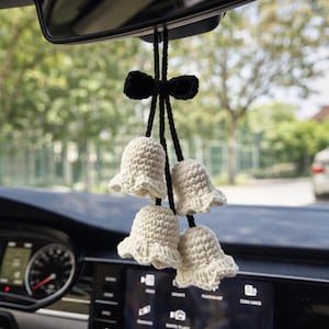 Pode incluir: Um amuleto de carro feito à mão com quatro ornamentos de crochê em forma de sino creme, suspensos por um laço e cordão pretos. O amuleto está pendurado no espelho retrovisor de um carro, adicionando um toque decorativo artesanal.