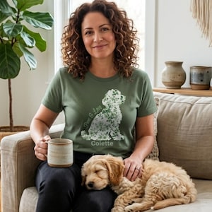 Könnte beinhalten: Ein olivgrünes Kurzarm-T-Shirt mit einer weißen, floralen Silhouette eines sitzenden Hundes. Die Grafik enthält den Schriftzug Paws & Love in Schreibschrift und den Namen Colette in einer serifenlosen Schrift unter der Hundeillustration.
