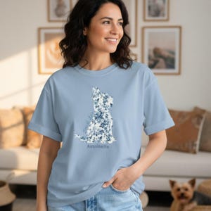 Puede incluir: Una camiseta de manga corta de color azul claro con un gráfico de la silueta de un perro relleno de un estampado floral azul y blanco. El texto "Antoinette" está impreso en una fuente azul oscuro justo debajo del gráfico floral en el pecho.