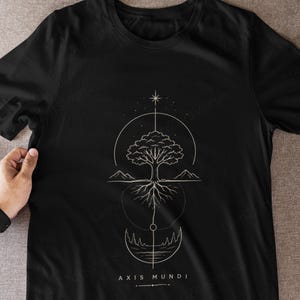 May include: A black short-sleeved t-shirt featuring a white line art graphic of a tree with visible roots, mountains, and a star. The design is enclosed in circular geometric elements and includes the text AXIS MUNDI printed below the illustration.