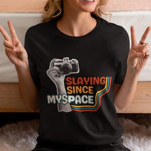 May include: A black short-sleeved t-shirt featuring a graphic print of a hand holding a vintage camera. The text reads "SLAYING SINCE MYSPACE" in orange and white, accompanied by three curved lines in orange, teal, and red colors.