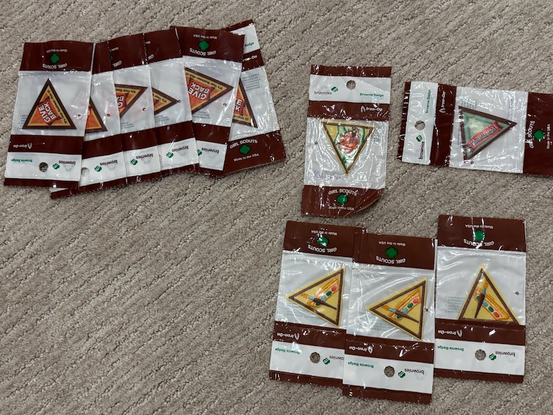 Pu&ograve; includere: Diversi distintivi Girl Scout Brownie in confezioni di plastica trasparente. I distintivi sono triangolari con vari disegni e colori, tra cui un distintivo con la scritta "Give Back". La confezione ha dettagli marroni e bianchi.