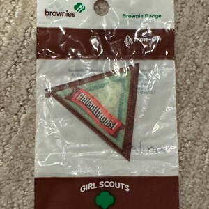 Pu&ograve; includere: Un distintivo Brownie in una confezione di plastica trasparente. Il distintivo &egrave; triangolare con uno sfondo verde e uno striscione rosso che dice "Philanthropist". La confezione riporta le parole "Brownies" e "Girl Scouts".