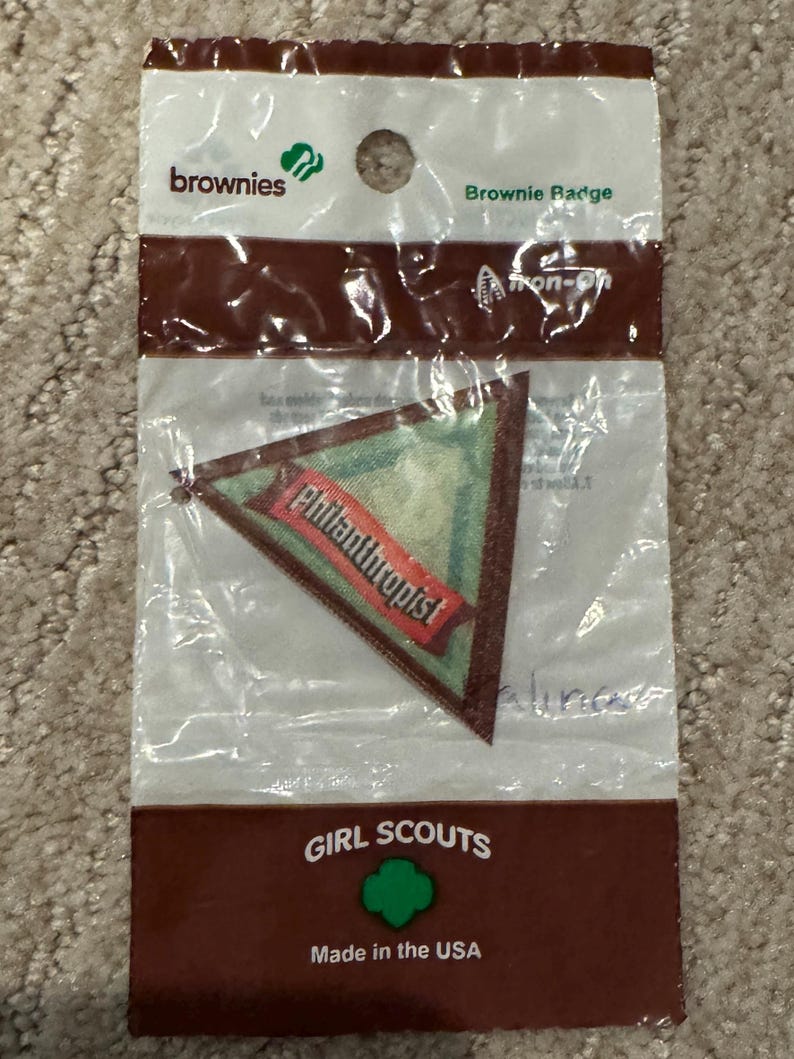 Pu&ograve; includere: Un distintivo Brownie in una confezione di plastica trasparente. Il distintivo &egrave; triangolare con uno sfondo verde e uno striscione rosso che dice "Philanthropist". La confezione riporta le parole "Brownies" e "Girl Scouts".