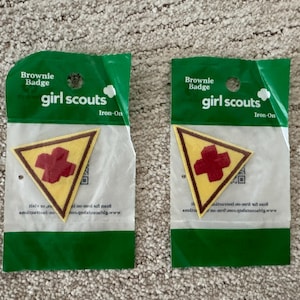 Pu&ograve; includere: Due toppe termoadesive Brownie Girl Scout. Ogni toppa &egrave; un triangolo giallo con un bordo marrone e una croce rossa al centro. Le toppe sono confezionate in plastica verde e bianca con le scritte "Brownie Badge" e "girl scouts".