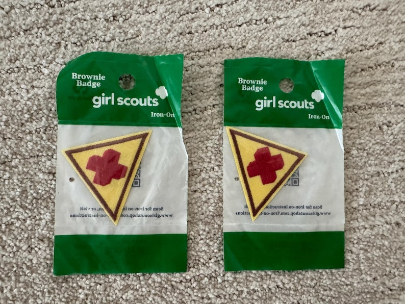 Pu&ograve; includere: Due toppe termoadesive Brownie Girl Scout. Ogni toppa &egrave; un triangolo giallo con un bordo marrone e una croce rossa al centro. Le toppe sono confezionate in plastica verde e bianca con le scritte "Brownie Badge" e "girl scouts".
