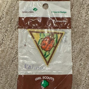 Pu&ograve; includere: Un distintivo Brownie in una confezione di plastica trasparente. La toppa triangolare presenta un disegno di coccinella su sfondo giallo con accenti verdi. La confezione recita "Brownies", "Brownie Badge" e "Girl Scouts Made in the USA."