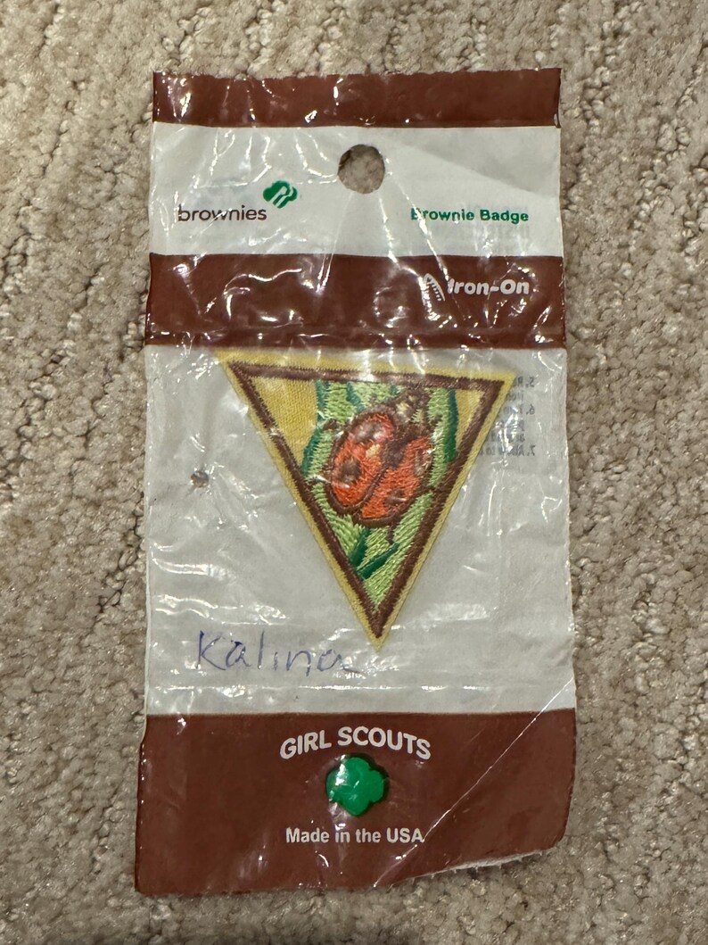 Pu&ograve; includere: Un distintivo Brownie in una confezione di plastica trasparente. La toppa triangolare presenta un disegno di coccinella su sfondo giallo con accenti verdi. La confezione recita "Brownies", "Brownie Badge" e "Girl Scouts Made in the USA."