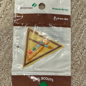 Pu&ograve; includere: Una toppa termoadesiva Brownie Badge in una confezione di plastica trasparente. La toppa triangolare ha un bordo giallo e un disegno ricamato con elementi marroni, blu, verdi e rossi. La confezione include il testo "brownies", "Brownie Badge", "Iron-On", "GIRL SCOUTS" e "Made in the USA".