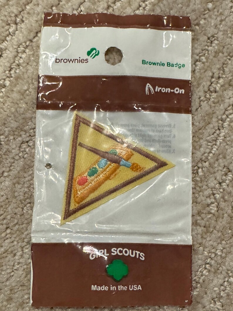 Pu&ograve; includere: Una toppa termoadesiva Brownie Badge in una confezione di plastica trasparente. La toppa triangolare ha un bordo giallo e un disegno ricamato con elementi marroni, blu, verdi e rossi. La confezione include il testo "brownies", "Brownie Badge", "Iron-On", "GIRL SCOUTS" e "Made in the USA".