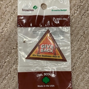 Pu&ograve; includere: Una confezione sigillata contenente una toppa termoadesiva Brownie Badge. La toppa triangolare presenta le parole "GIVE BACK" e "It's Your Business-Run It!". La confezione &egrave; marrone e bianca, con il logo delle Girl Scouts e la scritta "Made in the USA".