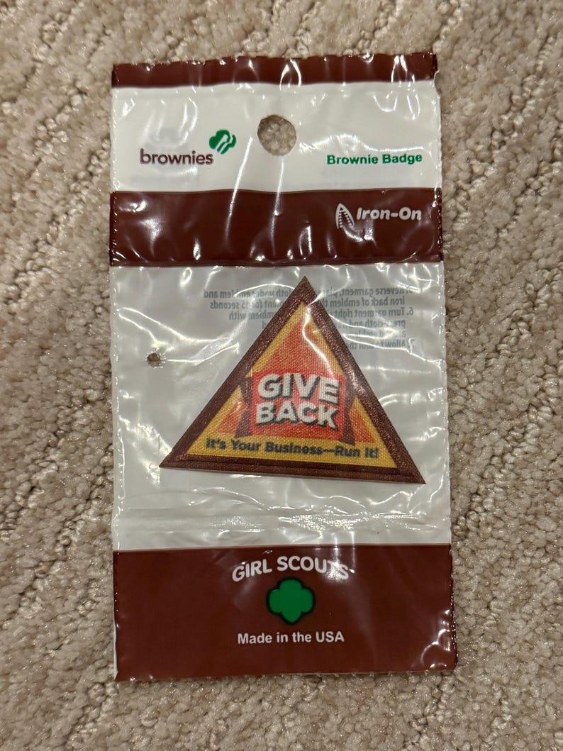 Pu&ograve; includere: Una confezione sigillata contenente una toppa termoadesiva Brownie Badge. La toppa triangolare presenta le parole "GIVE BACK" e "It's Your Business-Run It!". La confezione &egrave; marrone e bianca, con il logo delle Girl Scouts e la scritta "Made in the USA".