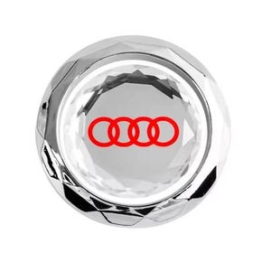 May include: A faceted, clear crystal car accessory with a red Audi logo. The logo features four interlocking rings. The accessory has a reflective, polished surface and a round shape.