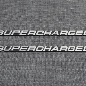 May include: Two silver-colored "SUPERCHARGED" emblems with a black background. The text is in a bold, sans-serif font. The emblems are displayed on a textured gray surface.