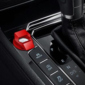 May include: A red, geometric car start button cover with the word "START" visible. The cover is set in a black car console, next to the gear shift and other control buttons.