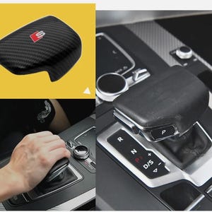 Op de afbeelding: Zwarte versnellingspookafdekking van koolstofvezel met een rood en zilver embleem. De afbeelding toont de afdekking op een gele achtergrond en geïnstalleerd in een auto. De versnellingspook is ook zichtbaar.