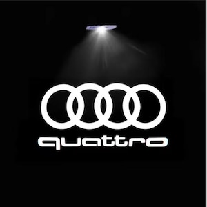 以下が含まれることがあります： 黒い背景に、4つのリングが組み合わさった白いアウディのロゴと、その下に「quattro」の文字。上部からスポットライトがロゴを照らしています。