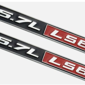 Puede incluir: Dos emblemas automotrices negros y rojos. La parte superior del emblema dice "5.7L" en letras blancas. La sección inferior derecha dice "LS6" en letras blancas. Los emblemas son rectangulares.