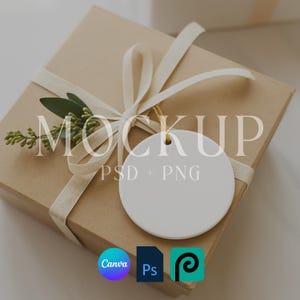 Könnte beinhalten: Eine quadratische, braune Geschenkbox, gebunden mit einem cremefarbenen Band und einem Zweig Grün. Ein weißer, runder Anhänger hängt am Band. Das Wort "MOCKUP" ist über das Bild gelegt, zusammen mit "PSD + PNG" und den Logos von Canva, Photoshop und PicsArt.
