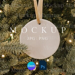 以下が含まれることがあります： ベージュのリボンで吊るされた、"MOCKUP"の文字がプリントされた白くて丸いクリスマスオーナメント。オーナメントは、ライトの付いたクリスマスツリーを背景に配置されています。"JPG, PNG"の文字も表示されています。