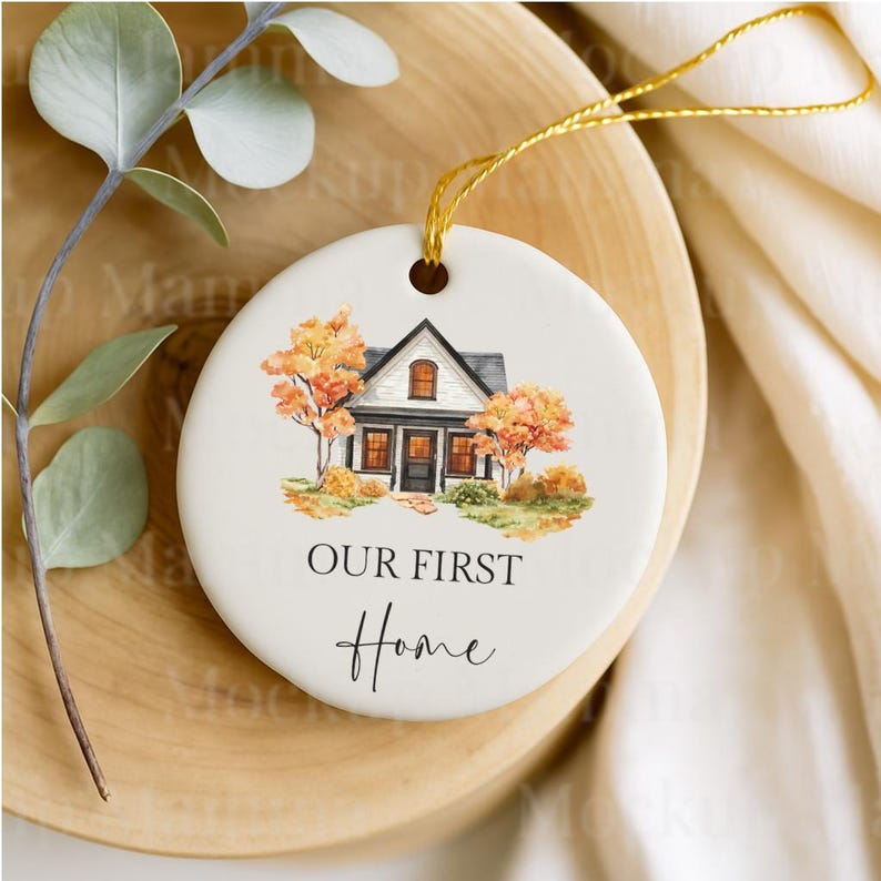 May include: Round ceramic ornament with a watercolor illustration of a house with fall foliage. The text "OUR FIRST Home" is written below the house. The ornament has a gold string for hanging.