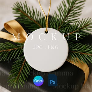 以下が含まれることがあります： 金色の紐が付いた、テキスト「MOCKUP」と「JPG . PNG」が表示された白い丸いオーナメント。オーナメントは、金色のリボンと常緑樹の小枝が付いた黒いギフトボックスの上に置かれています。CanvaとPsのロゴが表示されています。