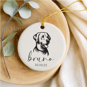 May include: A round, white ceramic ornament featuring a black and white illustration of a dog's head. The name "bruno" is written in cursive below the dog, with the date "10.10.25" beneath the name. A gold string is attached for hanging.