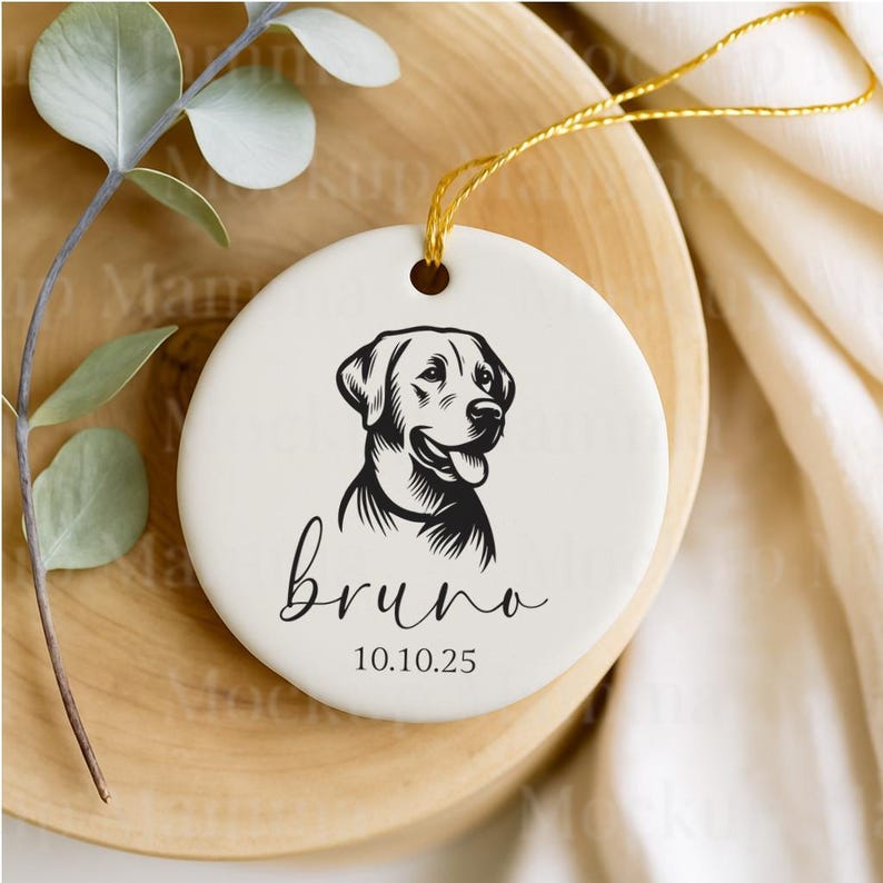 May include: A round, white ceramic ornament featuring a black and white illustration of a dog's head. The name "bruno" is written in cursive below the dog, with the date "10.10.25" beneath the name. A gold string is attached for hanging.