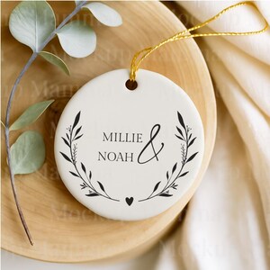 May include: A white ceramic ornament with the names "Millie & Noah" in black script, framed by a black floral wreath. A gold string is attached for hanging. The ornament is on a wooden surface.